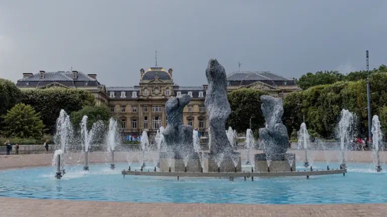 Fontaine monumentale au milieu d'un parc urbain verdoyant avec architecture classique en arrière-plan.