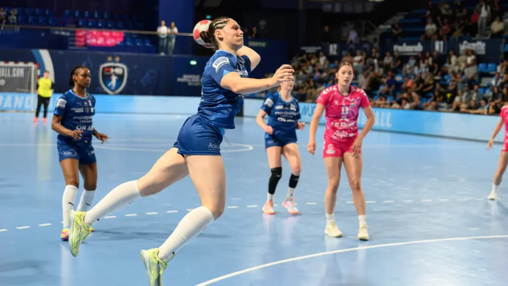 Une handballeuse en maillot bleu effectue un tir en suspension au-dessus du terrain.