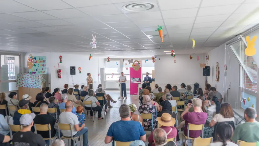 Une salle remplie de personnes assises écoutant trois intervenants debout devant un panneau rose.