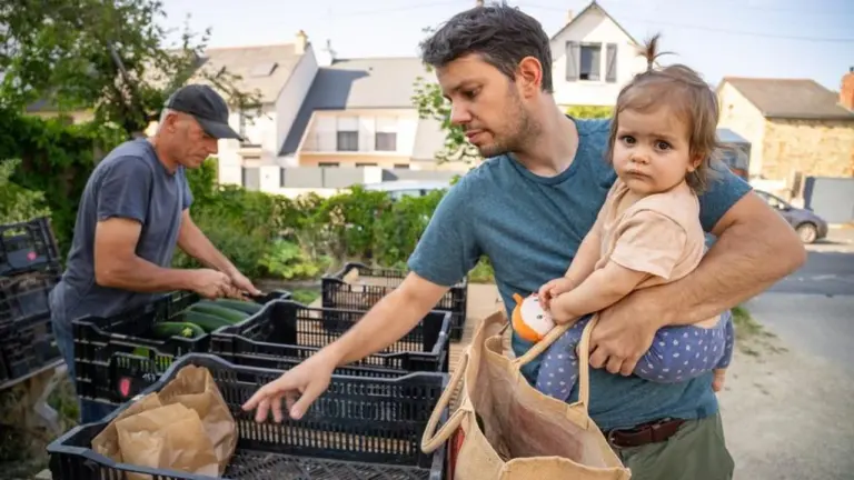 Un père porte son bébé tandis qu'un homme trie des courgettes dans des cagettes.