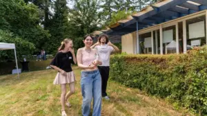 Trois jeunes adultes souriants marchent dans un jardin ensoleillé devant une maison moderne.