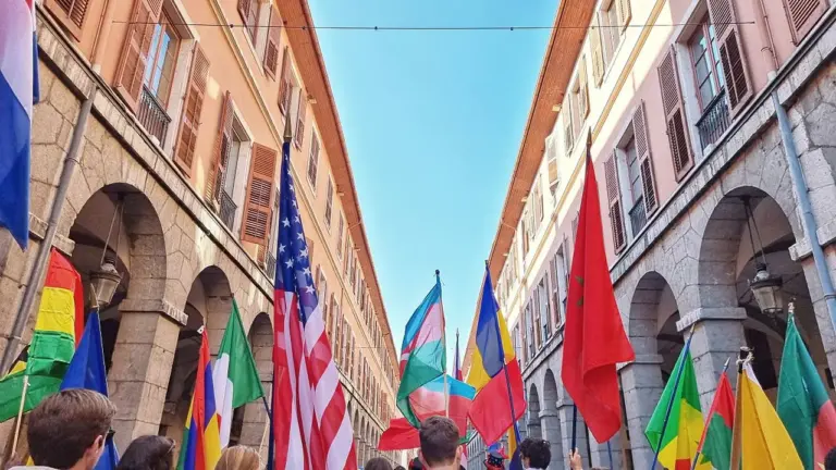 Une foule porte divers drapeaux nationaux dans une rue bordée de bâtiments historiques classiques.