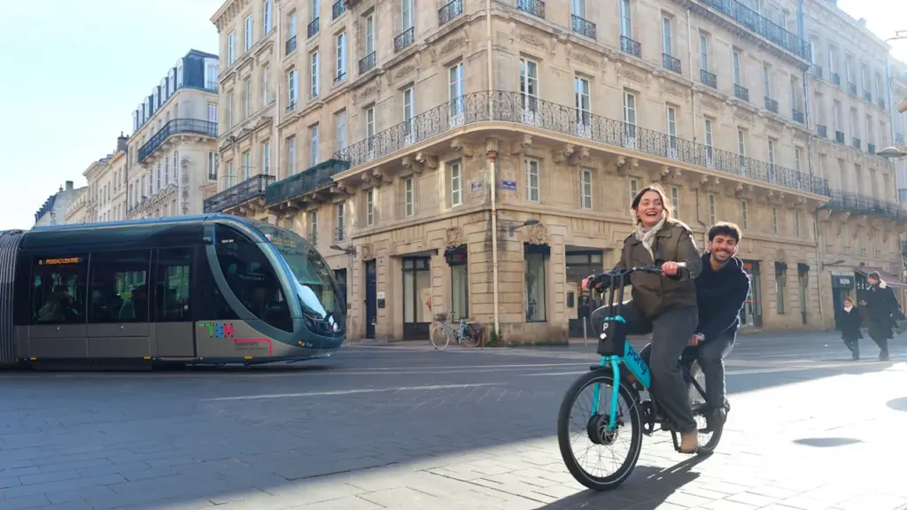 Deux personnes sur un vélo électrique à Bordeaux avec un tramway en arrière-plan.
