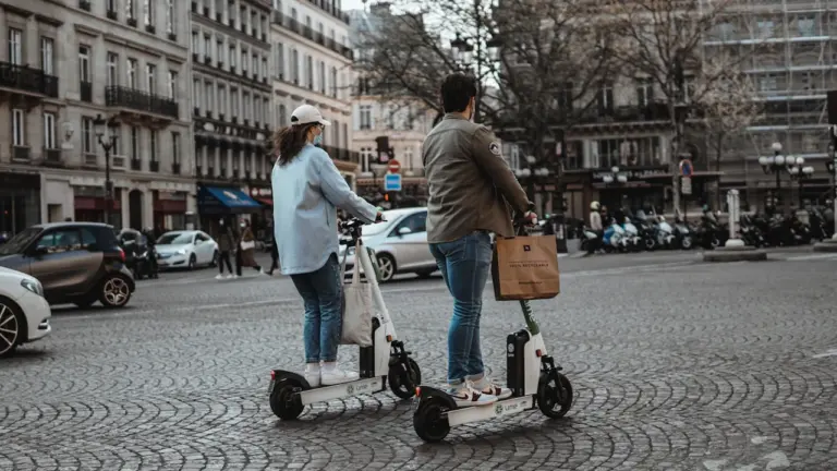 Deux personnes utilisent des trottinettes électriques sur une avenue pavée à Paris.