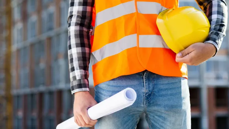 Un ouvrier portant un gilet orange et un casque jaune tient des plans architecturaux enroulés.