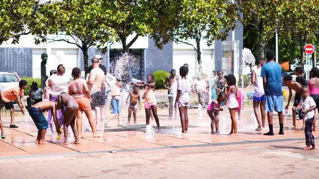 Un groupe d'enfants et d'adultes s'amuse dans des jets d'eau sur une place urbaine.