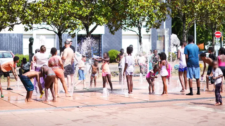 Un groupe d'enfants et d'adultes s'amuse dans des jets d'eau sur une place urbaine.
