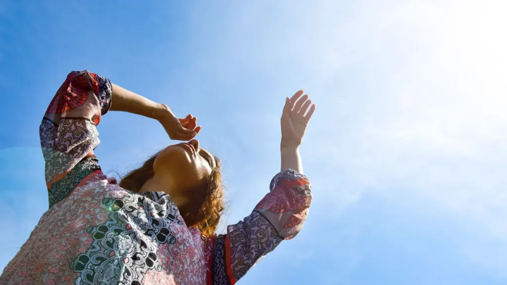 Une femme aux bras levés regarde vers le ciel bleu sous un soleil radieux.