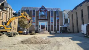 Une pelleteuse jaune travaille devant un bâtiment administratif sous un ciel bleu dégagé.