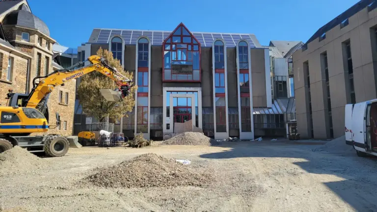 Une pelleteuse jaune travaille devant un bâtiment administratif sous un ciel bleu dégagé.