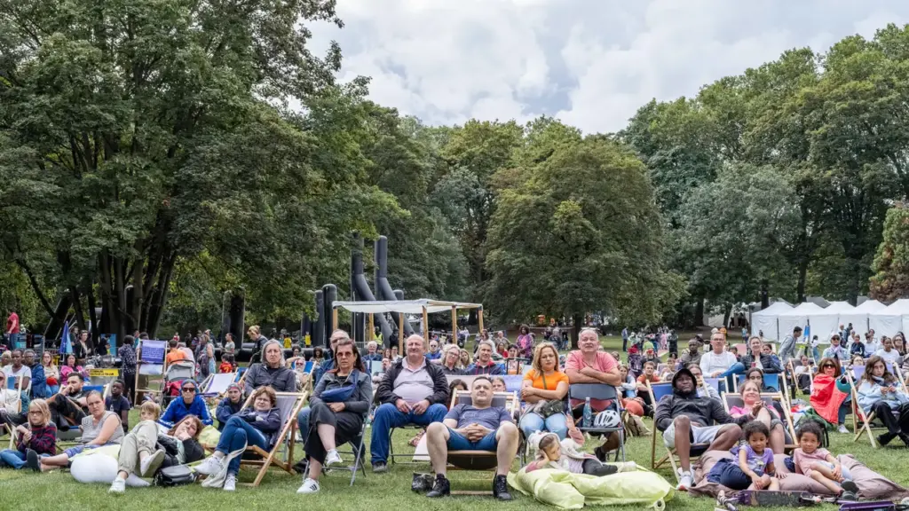 Un public diversifié assis sur des chaises longues dans un parc arboré et verdoyant.