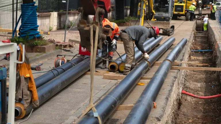 Ouvriers travaillant sur des canalisations noires près d'une tranchée et d'un engin de chantier.