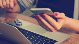 Une femme tient un smartphone devant un ordinateur portable posé sur une table en bois.