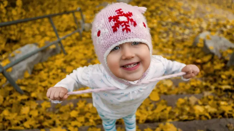 Une jeune enfant souriante porte un bonnet rose dans des feuilles d'automne jaunies.